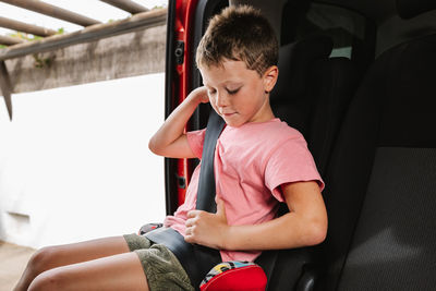 Unemotional boy in casual clothes buckling up while sitting on seat inside of car on back seat of car during road trip