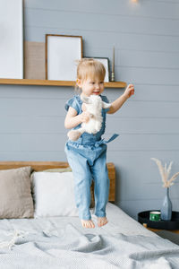 Cute two-year-old girl in a denim jumpsuit with a soft toy in her hands jumps on the bed person