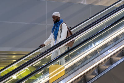 African american passenger man with suitcase stands on escalator, holds handrail in airport terminal