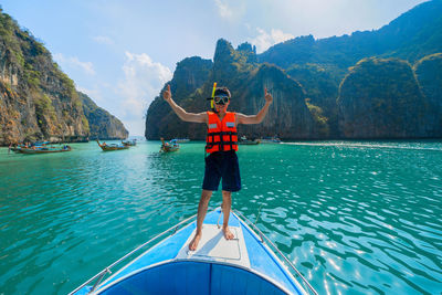 Full length of man wearing life jacket while standing on sailboat in sea against mountains