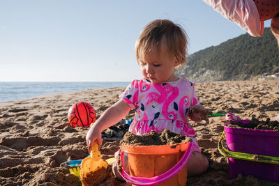 Baby girl in pink dress sitting in a sand with sand toys at the beach