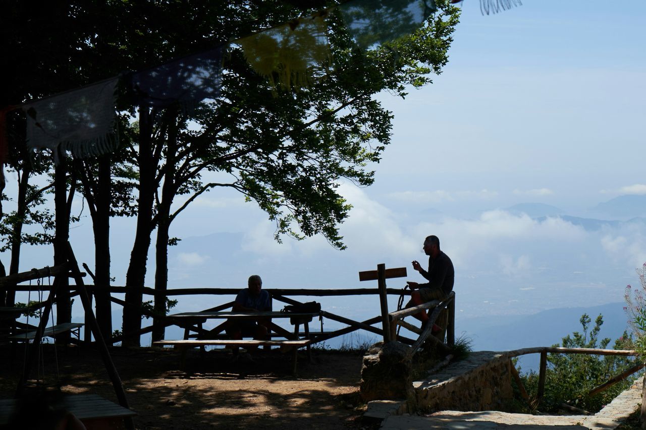 sky, silhouette, built structure, sitting, sunlight, men, low angle view, railing, leisure activity, architecture, tree, bench, outdoors, nature, lifestyles, wood - material, relaxation, day