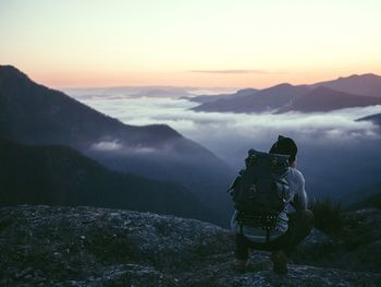 Full length rear view of hiker crouching on cliff by cloudscape and mountains at morning