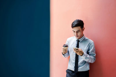 Young man holding coffee while standing against wall