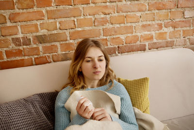 Portrait of beautiful young woman sitting on sofa against brick wall