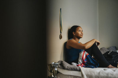 Teenage girl day dreaming sitting on bed against wall in bedroom at home