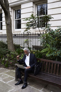 Senior businessman sitting on bench outdoors working on laptop