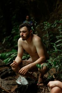 Young man looking away while sitting on rock in forest