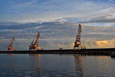 Metallic cranes at commercial dock at harbor against cloudy sky