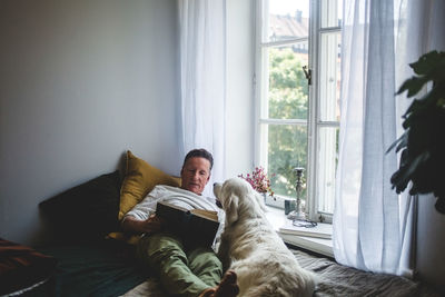 Senior man lying with dog while reading book on bed by window at home
