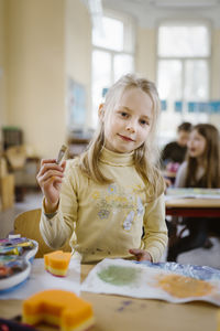Portrait of female blond student holding paintbrush in classroom