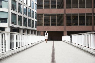 Mature businesswoman talking through smart phone while walking on footbridge against office building at downtown district