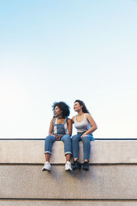 Female friends sitting on retaining wall against clear sky