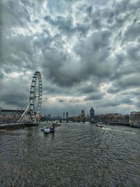 View of ferris wheel by river against cloudy sky