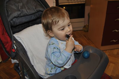 Cute boy licking spoon while sitting on baby stroller at home