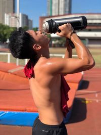 Side view of shirtless sweaty man drinking water while standing on sports track