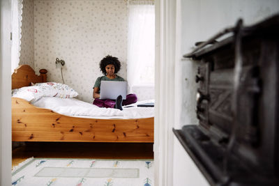Young woman using laptop while relaxing on bed at home seen through doorway