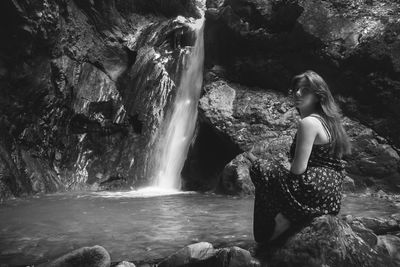 Portrait of young woman sitting by waterfall on rock