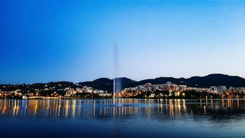 Illuminated city by lake against clear blue sky