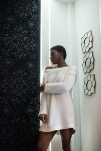 African american female in white trendy dress standing near wall in room and looking away