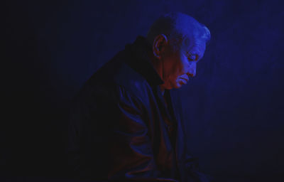 Depressed lonely senior man illuminated with blue neon light against black background