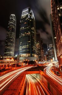 Light trails on road amidst illuminated buildings against sky at night