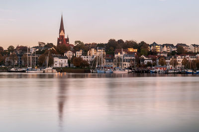 Blurred motion of river by buildings in city against sky during sunset