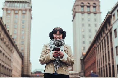 Smiling young woman wearing warm clothing while using mobile phone against buildings in city