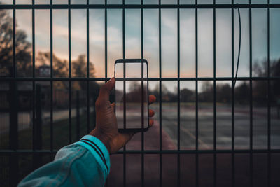 Cropped hand using smart phone against fence during sunset