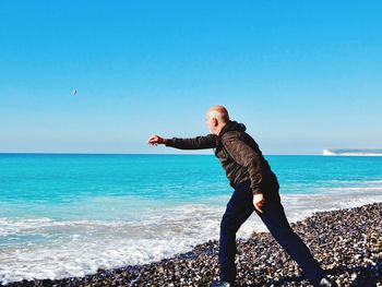 Side view of man throwing pebble in sea against blue sky