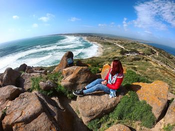 Woman looking at sea while sitting on rocks against sky