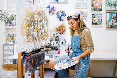 Side view of female artist sitting near easel and drawing colorful picture with paints while working in creative workshop