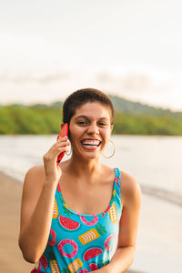 Cheerful young latin american woman with short hairstyle talking on mobile phone while standing near waving sea in summer day