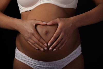 Midsection of pregnant woman making heart shape with hands on her belly against black background
