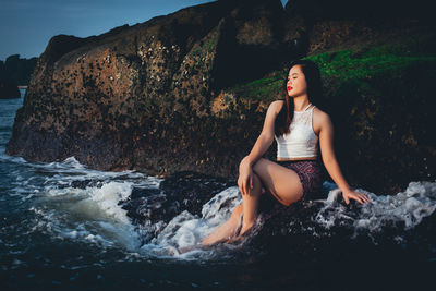 Young woman sitting on rock by sea against sky