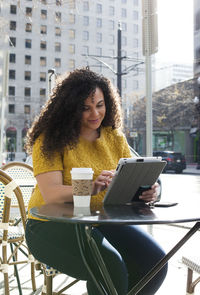 Woman using tablet computer while sitting on chair at sidewalk cafe in city