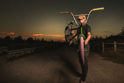 Portrait of young man carrying bicycle while standing on dirt road against sky during sunset