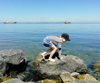 Cheerful girl crouching on rock by sea against sky