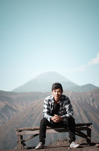 Portrait of young man sitting on bench against mountains during sunny day