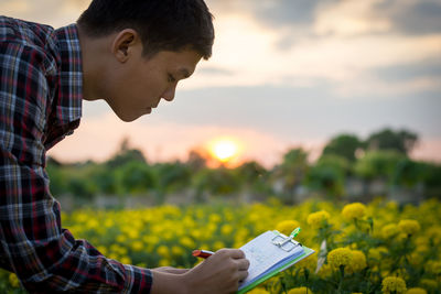 Farmer writing on clipboard while examining flowers on field during sunset