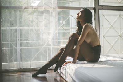 Side view of young woman sitting on bed by window
