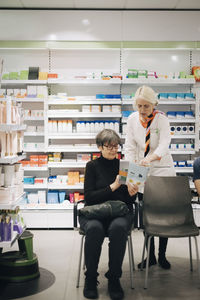 Full length of senior customer sitting on chair showing magazine to female owner against rack at pharmacy store