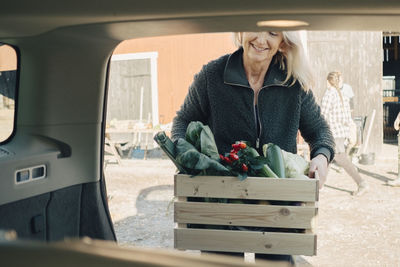 Mature woman loading crate full of organic vegetables in car trunk