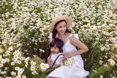 Woman in a white dress with her son  in a chamomile field at sunset in summer