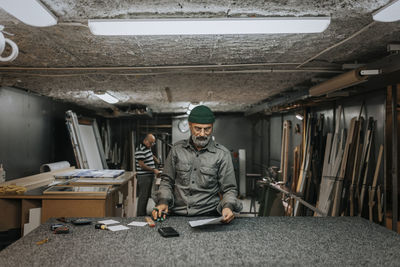 Male owner reading blueprint at workbench with colleague working in background