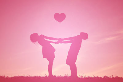 Boy and girl holding hands while standing below heart shape against sky during sunset