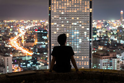 Rear view of man sitting against illuminated buildings at night