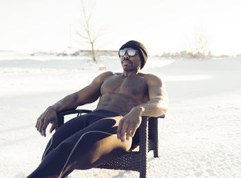 Shirtless male athlete looking away while relaxing on chair in snow