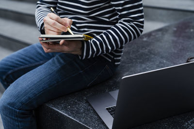 Midsection of man writing in book while using laptop