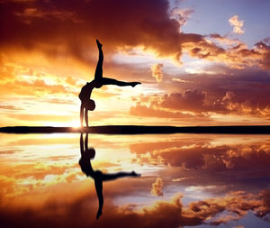 Silhouette woman performing handstand with reflection on lake against cloudy sky during sunset